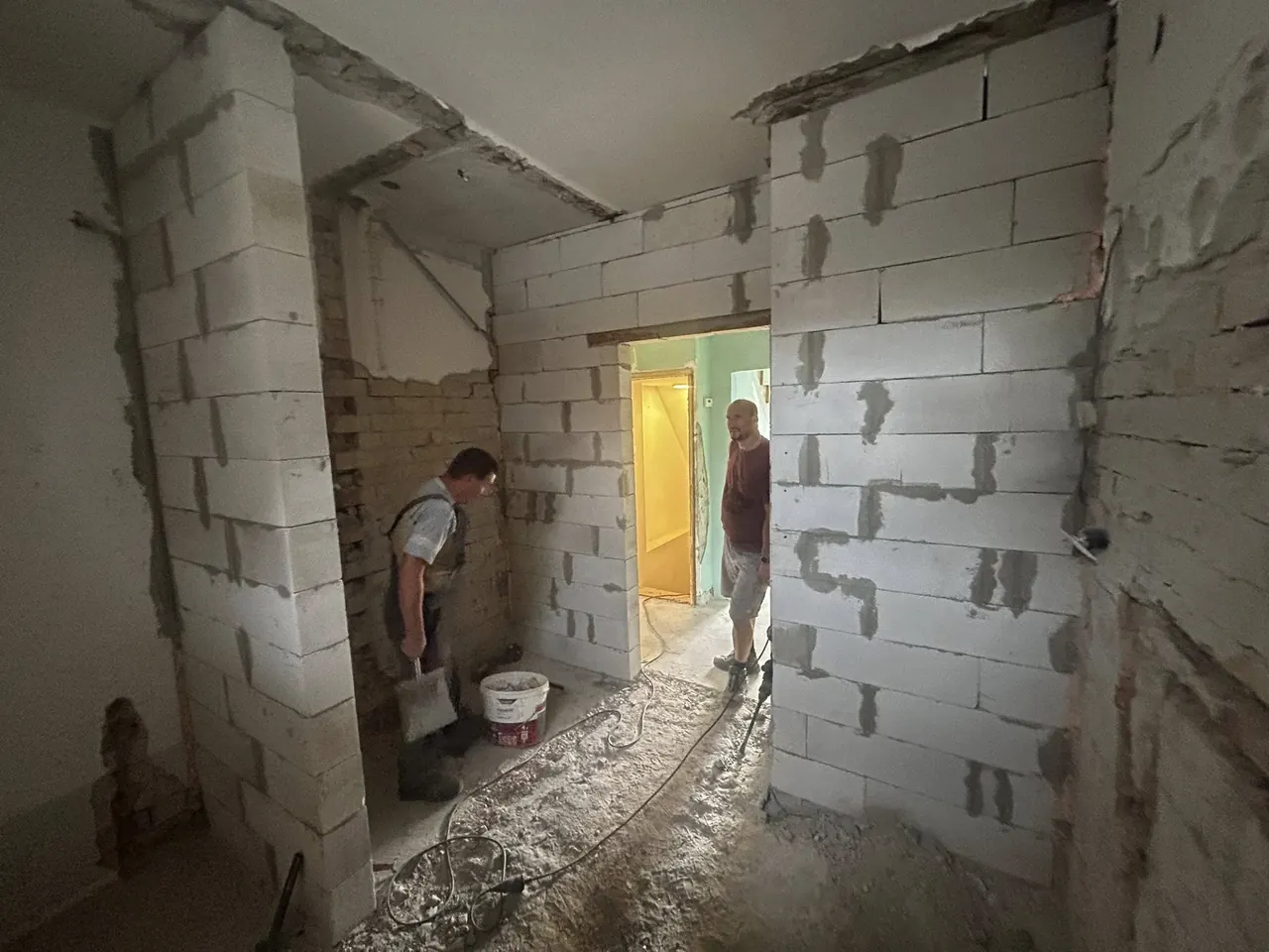 Két munkás dolgozik egy építkezésen Győr városában, ahol a Farkas-Konstrukt Kft. generálkivitelezési projektje zajlik. Az egyik munkás falazó anyagot hordoz, míg a másik az elkészült falakat vizsgálja. A háttérben látható az építkezés előrehaladása, a még befejezésre váró térelválasztó falak és az építőanyagok.