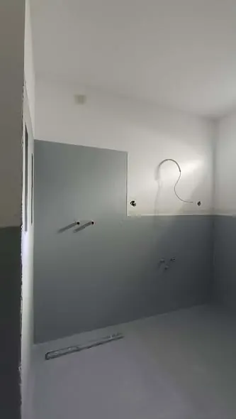 A képen egy frissen felújított helyiség látható, ahol a falak világosszürke színben vannak festve, és a padló sima, világos felületű. A falon látható a vízvezetékek előkészítése, valamint egy világítótest is. Ez a generálkivitelezés Győrben, a Farkas-Konstrukt Kft. által végzett munkáját tükrözi, amely a modern belső terek kialakítására összpontosít.