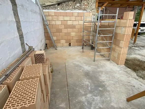 A képen látható egy építkezési terület, ahol a falazás folyamatban van. A háttérben látható a már elkészült fal, míg az előtérben téglák találhatók, amelyek készen állnak a felhasználásra. Az építkezés Győr városában zajlik, a Farkas-Konstrukt Kft. generálkivitelezési szolgáltatásának keretében. A területen állványok is láthatók, amelyek segítik a munkát.