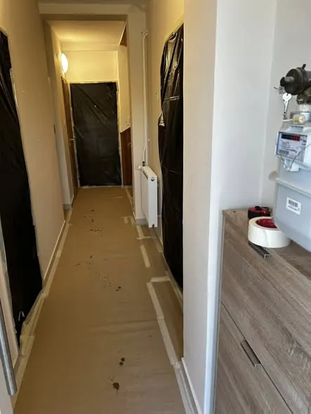 A képen egy szűk folyosó látható, amelyen a falakat fekete fóliával borították, valószínűleg felújítás vagy festés céljából. A padlón védőfólia található, és a falakon radiátorok láthatók. A folyosó végén egy ajtó, míg a fal melletti bútor egy komód. A Farkas-Konstrukt Kft. Generálkivitelezés szolgáltatásaival Győr városában várja ügyfeleit.