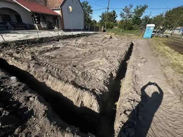 A képen egy frissen ásott árok látható, amely Győr városában található, és a Farkas-Konstrukt Kft. generálkivitelezési projektjéhez tartozik. Az árok szélei egyenletesen meg vannak formázva, a háttérben pedig egy épület körvonalai és néhány fa látható. A terület körül kerítés van, ami az építkezés biztonságát szolgálja. A napfény erősen világít, ami hangsúlyozza a terület részleteit.