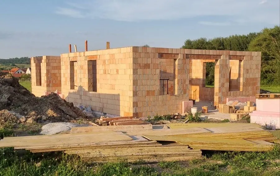 Győrben, a Farkas-Konstrukt Kft. generálkivitelezésének projektjén egy új épület áll, amely éppen építés alatt áll. Az épület falai téglából készülnek, és a környezetben láthatók az építkezéshez szükséges anyagok, mint a fakanalak és az építőanyagok. A napfény világítja meg a területet, amely zöldellő fákkal és dombokkal körülvett.