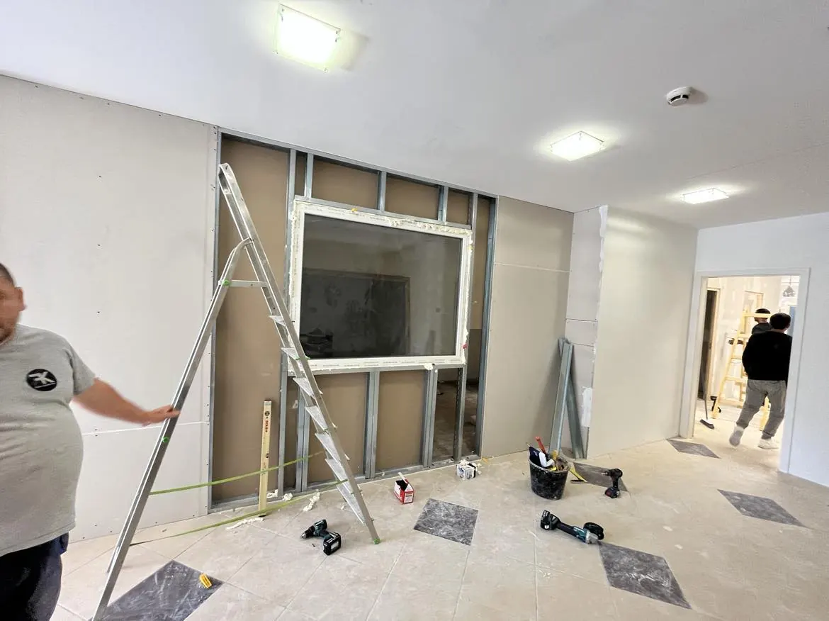 A képen egy generálkivitelezési munkafolyamat látható Győr városában. Az előtérben lévő létra mellett egy munkás áll, miközben a falon egy új nyílás kialakítása zajlik. A padlón szerszámok és anyagok találhatók, jelezve a felújítás folyamatát. Az építkezés során a falak és padló burkolata is látható, ami a Farkas-Konstrukt Kft. szakértelmét tükrözi.