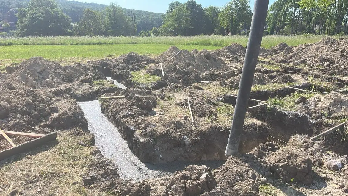 A képen egy építkezési terület látható, ahol a Farkas-Konstrukt Kft. Generálkivitelezés tevékenysége zajlik Csornán. A földmunkák során ásott alapok és kitöltött betoncsövek láthatók, amelyeket fák és zöld fű vesz körül. A terület rendezett, és jól látható, hogy a jövőbeli épület alapozása folyamatban van.