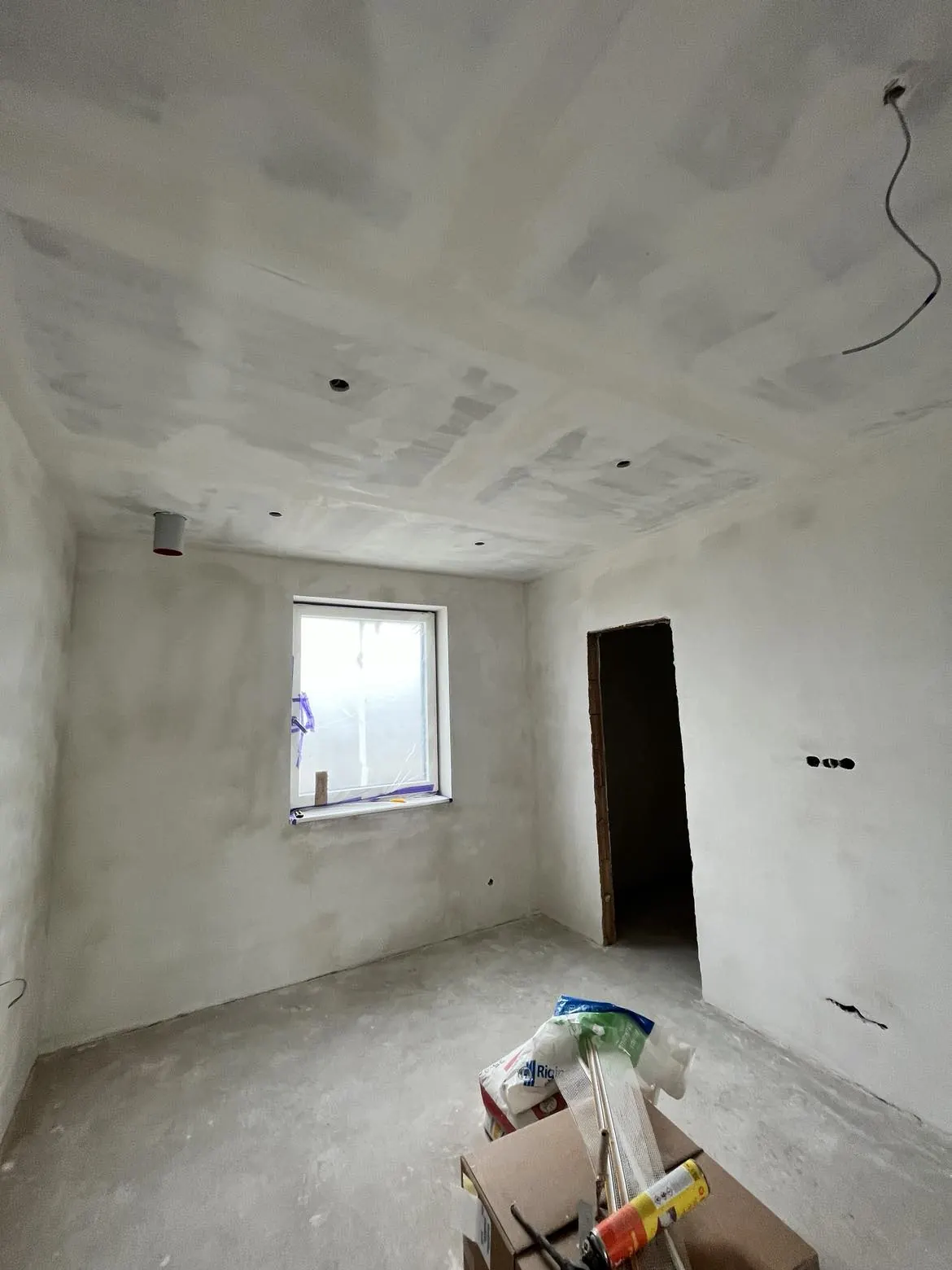 A képen egy frissen felújított szoba látható, amelynek falai világos színűre vannak meszelve, és a plafonon még láthatóak a festés előtti előkészítési munkák nyomai. Az ablak előtt természetes fény áramlik be, míg az ajtókeret nyitott, de az ajtó hiányzik. Az építkezési anyagok, mint például ragasztó és szigetelő anyagok, a szoba közepén találhatók. A Farkas-Konstrukt Kft. generálkivitelezésével készült felújítás a komáromi ingatlanban zajlik.