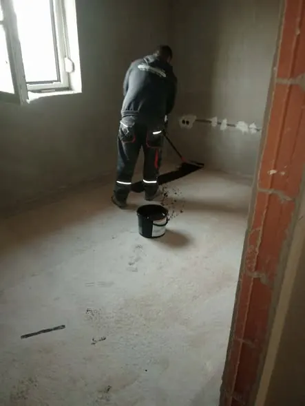 Egy munkás a Farkas-Konstrukt Kft. generálkivitelezési projektjén dolgozik Győr városában. A képen látható, ahogy a férfi simítja a frissen elkészített padlót, miközben egy vödör festék áll a közelben. A falak még befejezésre várnak, és a környezet ipari jellegű, amely a kivitelezési munkálatok folyamatát tükrözi.