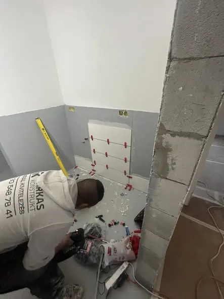 Egy férfi a Farkas-Konstrukt Kft. generálkivitelezési projektjén dolgozik Győr városában. A képen látható, amint a falat készíti elő, fektetési és burkolási munkákat végez, miközben különböző szerszámokat és anyagokat használ. A háttérben a falon látható a helyszín előkészítése, ami a precíziós munkát jelzi.