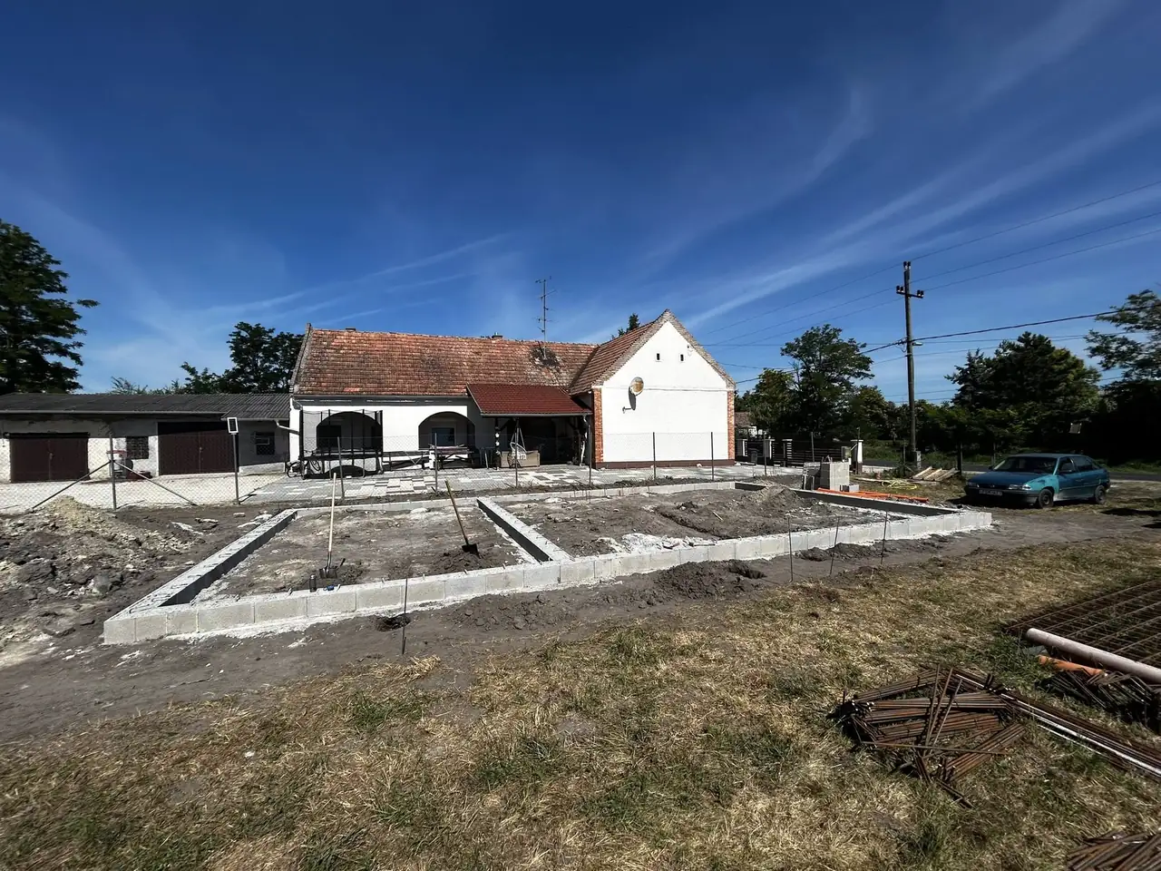 A képen egy építkezési terület látható Győrben, ahol a Farkas-Konstrukt Kft. generálkivitelezési munkálatai zajlanak. Az előtérben a frissen kialakított alapozás látható, amely körül zöld fű és néhány építőanyag hever. A háttérben egy fehér falú, piros tetős épület áll, amely felújítás alatt áll. Az ég kék és felhőtlen, ami ideális időjárási körülményeket teremt az építkezéshez.