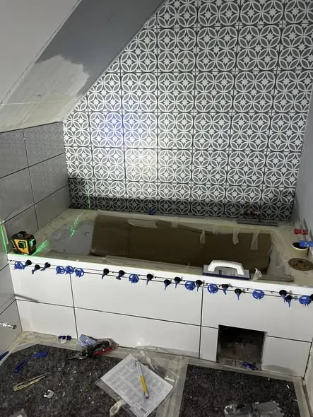 A képen egy fürdőszoba látható, ahol a csempe burkolása folyik. A falon modern, geometrikus mintázatú csempe található, míg a kád körüli terület még befejezésre vár. Az aljzatot burkoló anyagok és szerszámok szétszórva hevernek, jelezve a Generálkivitelezés folyamatát. A kép Győr városában készült, ahol a Farkas-Konstrukt Kft. szakemberei dolgoznak.