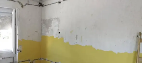 A képen egy győri generálkivitelezési projekt látható, ahol egy fal részben már le van festve sárga színűre, míg a többi rész fehér. A falon láthatók a festés előkészítésének jelei, például a csupasz részek és a tapétázott területek. A helyiség világos, természetes fényben úszik, ami hangsúlyozza a felújítás alatt álló teret. A Farkas-Konstrukt Kft. szakszerűen végzi a felújítást.