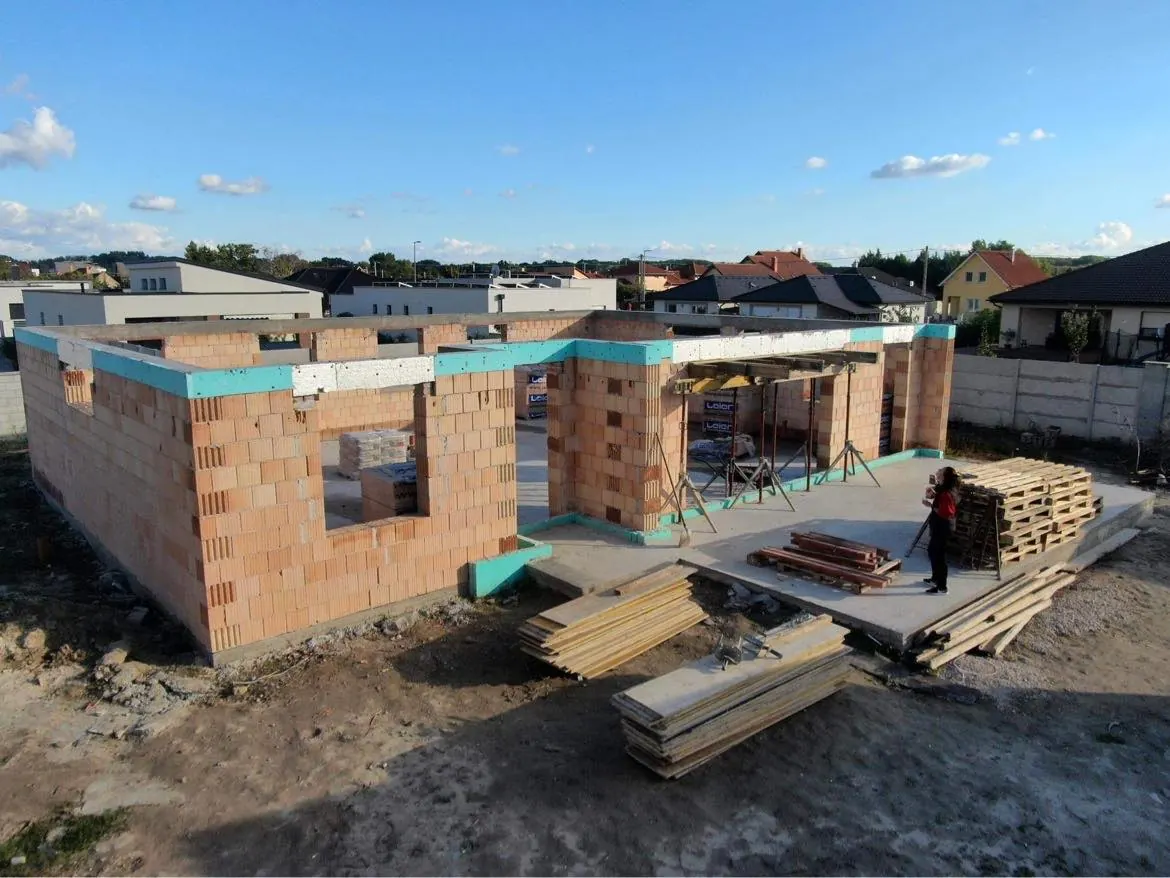 A képen egy építkezés látható Győr városában, ahol a Farkas-Konstrukt Kft. generálkivitelezési szolgáltatását végzi. A falazás folyamatban van, láthatóan téglaelemekből épül a ház szerkezete. Az épület körüli területen faanyagok és építőanyagok találhatók, míg az égen derült idő van. Az építkezés modern, és jól megtervezett.