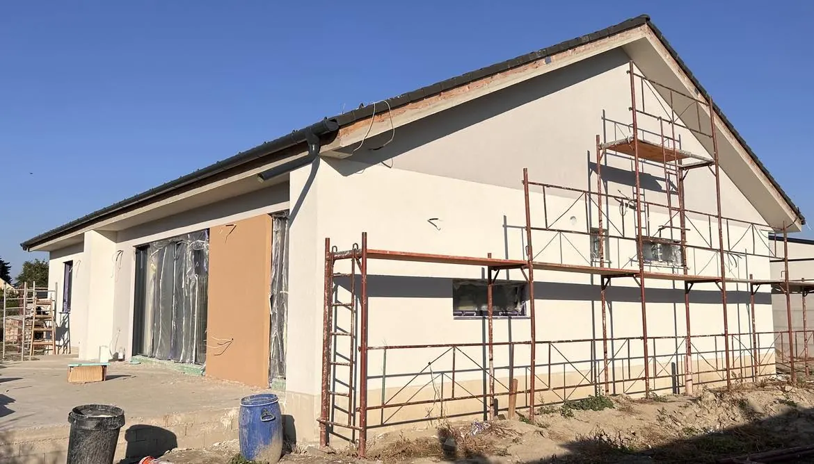 Győr városában, a Farkas-Konstrukt Kft. generálkivitelezési projektjén látható egy épülő ház, amelyen éppen a külső falak festése és befejezése zajlik. Az épület mellett állványzat található, amely segíti a kivitelezőket a magasabb részek elérésében. A környező terület rendezett, és az építkezési anyagok jól láthatóak.