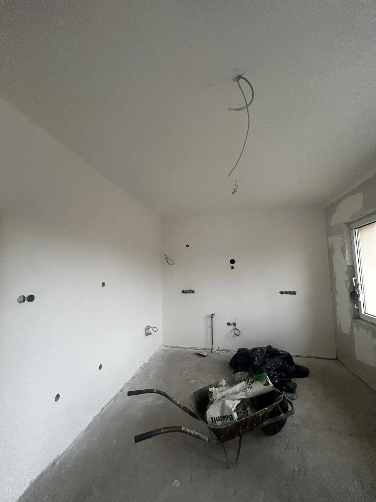 A képen egy üres, frissen kiépített helyiség látható, amelynek falai simára vakoltak, de még festésre várnak. A padlóbeton látható, és a falakon elektromos szerelvények helyének előkészítése zajlik. A sarokban egy talicskában építőanyagok találhatóak. A Farkas-Konstrukt Kft. generálkivitelezési szolgáltatása Győr városában zajlik, amely a modern építkezések igényeit szolgálja ki.