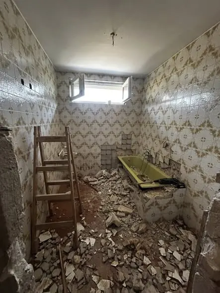 A képen egy felújítás alatt álló fürdőszoba látható, ahol a padlón törmelékek hevernek, és a falakon régi csempe található. A falak mintázata virágos, de a helyiség rendkívül rendezetlen. Az ablakok nyitva vannak, és egy létra áll a szobában, ami a felújítási munkálatokat jelzi. Ez a helyszín Kapuvárban található, ahol a Farkas-Konstrukt Kft. Generálkivitelezés szolgáltatásait végzi.