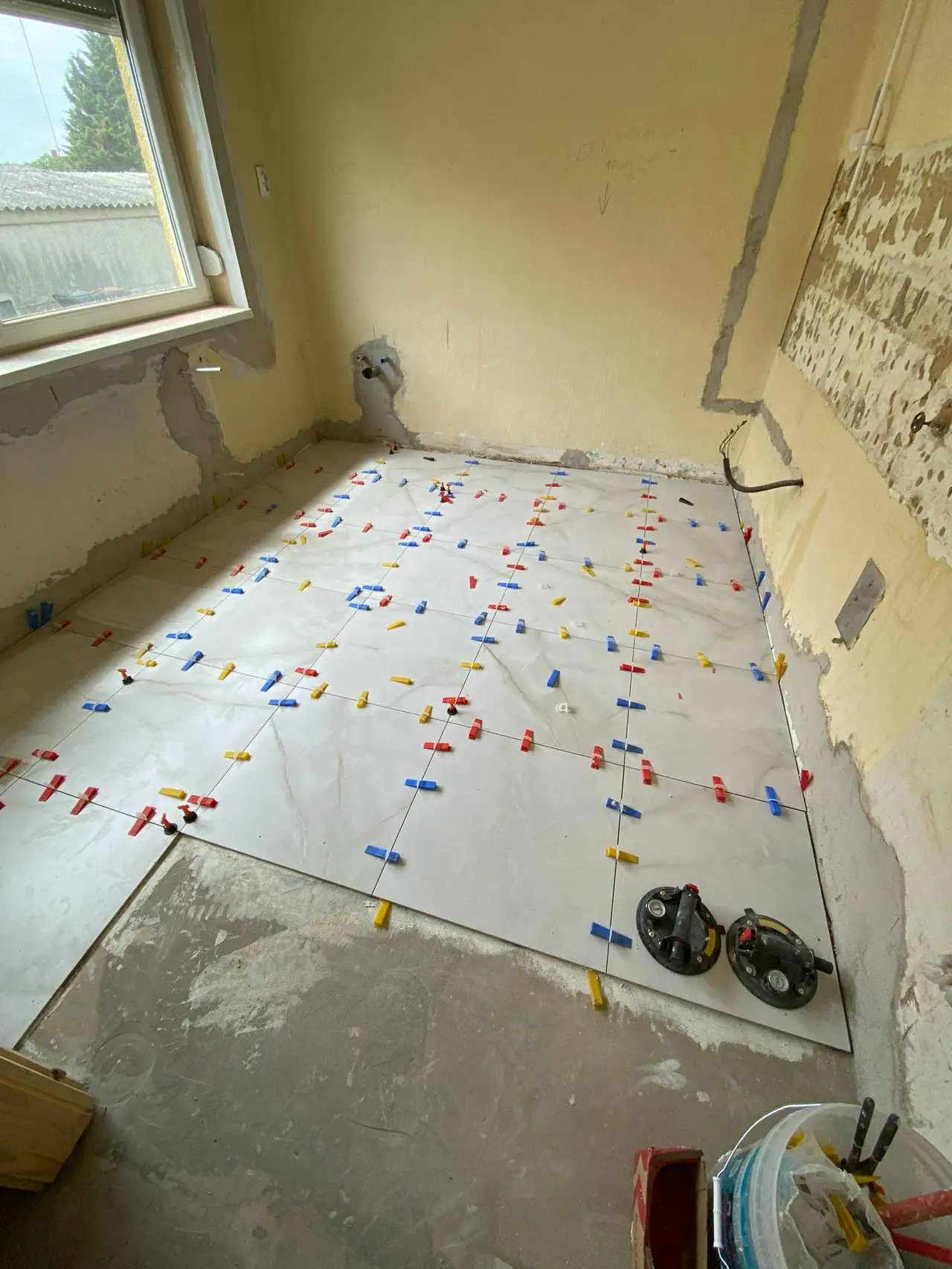 Az Oroszlányban található Farkas-Konstrukt Kft. generálkivitelezési projektje során egy belső tér burkolásának előkészítése látható. A falak még nem készültek el, de a padlón különböző színű, műanyag spacerek találhatók, amelyek a csempe egyenletes elhelyezését segítik elő. Az eszközök, mint például a csempevágó, a munkafolyamat során hasznosak lehetnek.