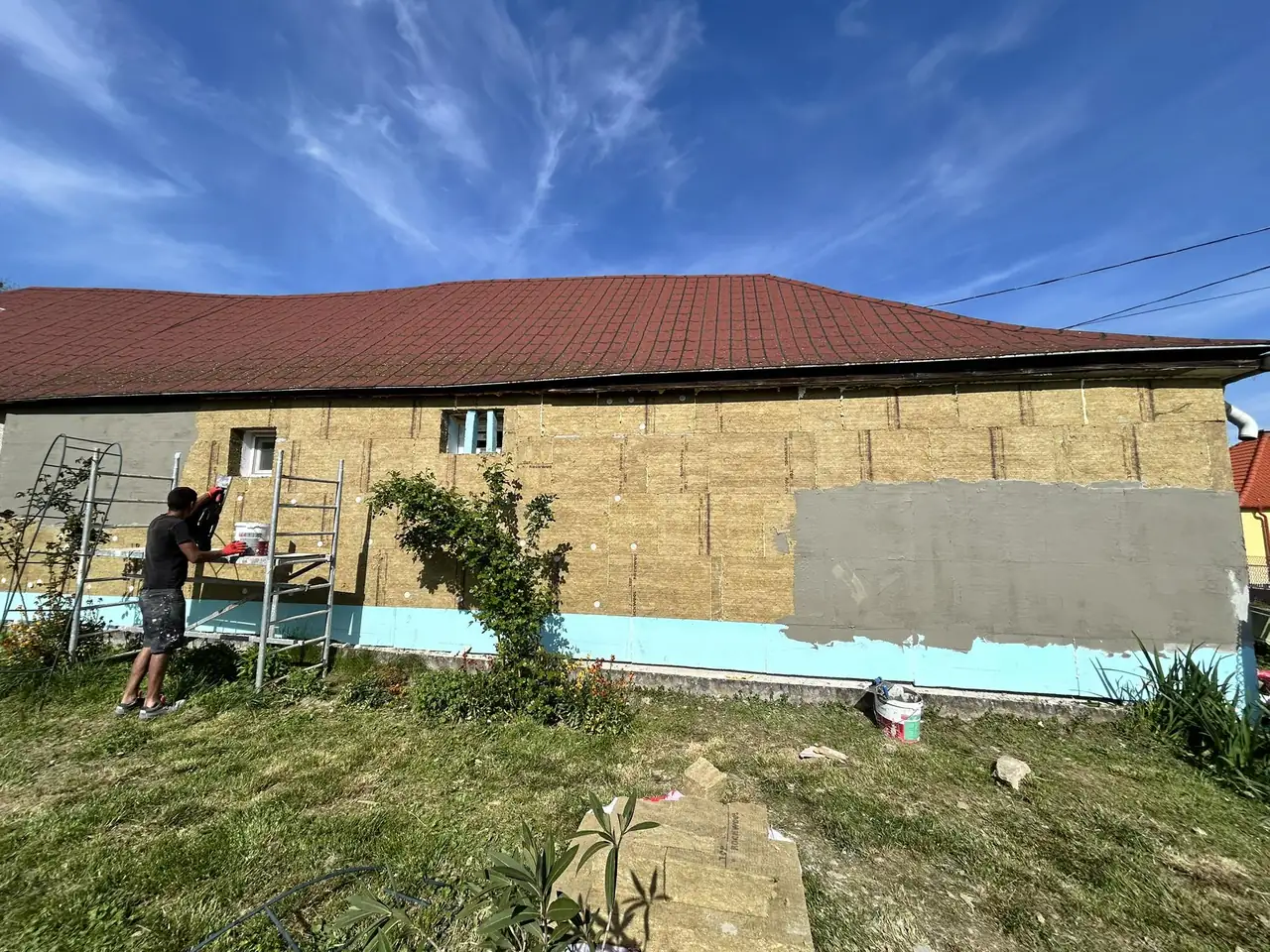 A képen egy ház felújítása látható Komáromban, ahol a Farkas-Konstrukt Kft. generálkivitelezési munkálatait végzik. Az épület oldalfala szigetelés alatt áll, a munkások állványon dolgoznak, míg a háttérben gyönyörű kék ég és fehér felhők láthatók. Az udvarban növények és festékes vödrök találhatók, ami a felújítás folyamatát tükrözi.