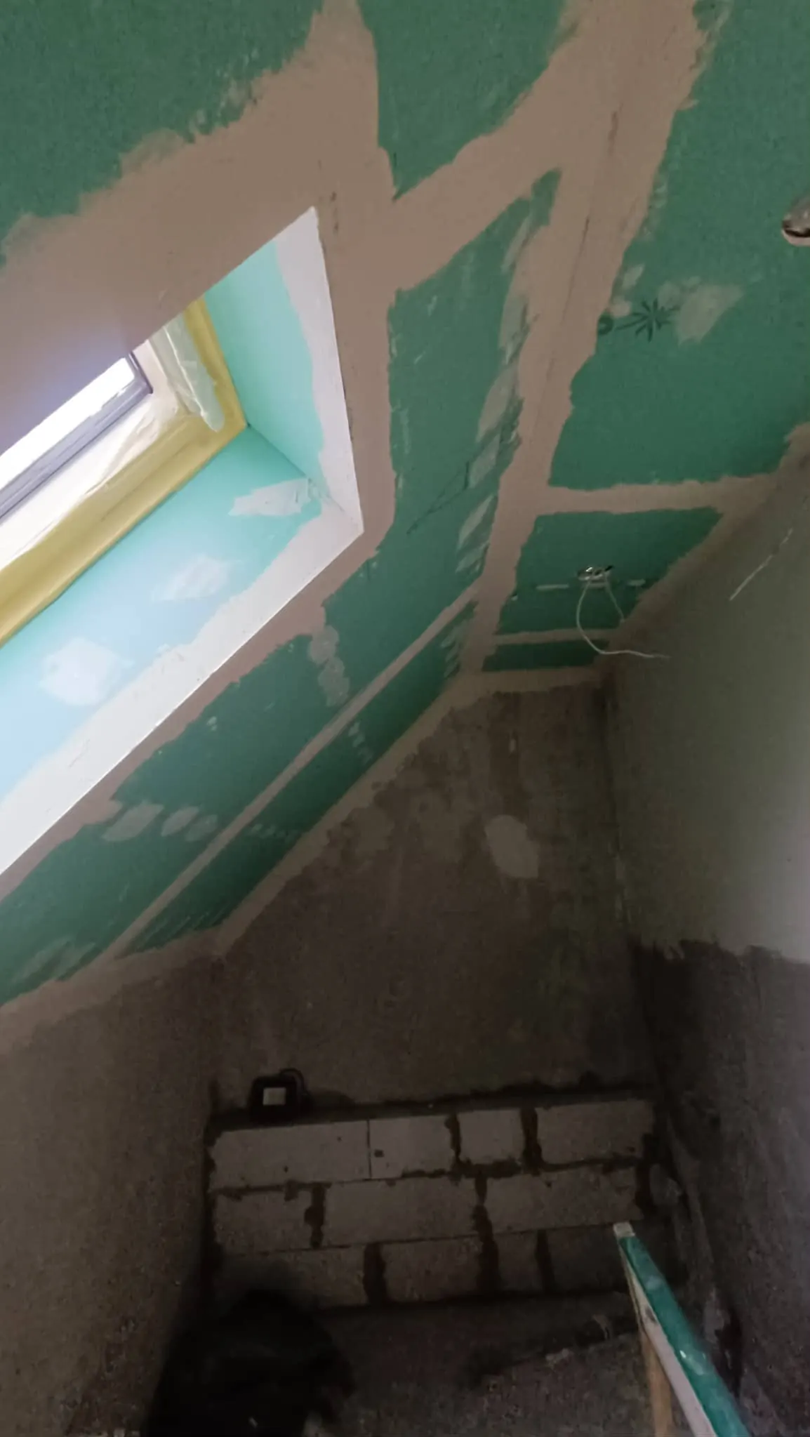 A képen egy építkezés alatt álló fürdőszoba látható, ahol a falakat zöld gipszkarton borítja. A szoba sarkában egy tetőablak található, amelyen keresztül természetes fény árad be. A padló még nem készült el, és az alapozás is látható. A Farkas-Konstrukt Kft. generálkivitelezési munkáit tükrözi, amely Győr városában zajlik.