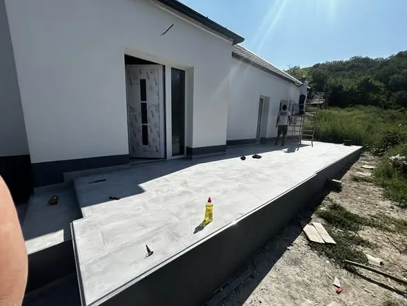 A képen egy frissen épült ház látható Győrben, ahol a Farkas-Konstrukt Kft. generálkivitelezési munkálatai zajlanak. Az épület előtt sima, világosszürke burkolat található, amelyet még nem véglegesítettek teljesen. A háttérben egy munkás látható, aki a kivitelezés részletein dolgozik. Az épület modern formavilága és a környező zöld terület harmonikus összképet alkot.