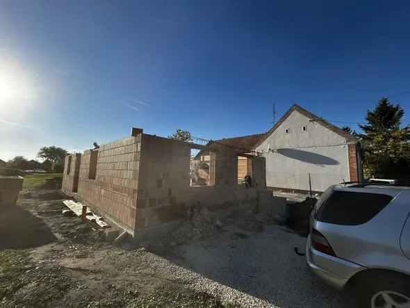 A képen a Farkas-Konstrukt Kft. generálkivitelezési munkálatai láthatók Csornán. Az építkezés során éppen egy új épület falainak felhúzása zajlik, a háttérben egy már meglévő ház áll. A napfény világítja meg a területet, ahol az építőanyagok és szerszámok is megtalálhatók, jelezve a folyamatban lévő munkát.