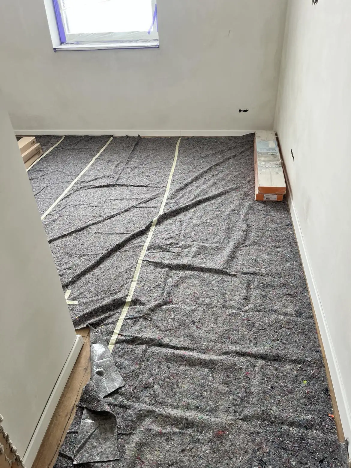 Újonnan felújított helyiség, ahol a padlóburkolás előkészítése zajlik. A falak világos színűek, a padlón fekete szőnyeg alátét található, mellette világos faanyagokkal. Az ablak nyitva van, természetes fény áramlik be. A Farkas-Konstrukt Kft. Generálkivitelezés szolgáltatásai Győr városában tükröződnek a precíz munkában.