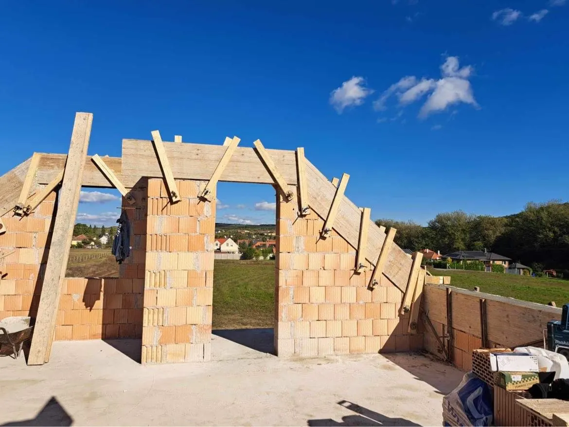 A képen egy építkezés látható Győrben, ahol a Farkas-Konstrukt Kft. generálkivitelezési munkálatai zajlanak. Az épület falai már láthatóan elkészültek, a téglák és a fa támaszok jól kivehetőek. A háttérben zöld dombok és házak látszanak, a kék égbolt pedig felhőkkel díszített. Az építkezés területe rendezett, a munka folytatódik.