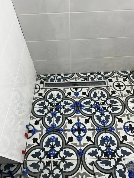 A képen a Farkas-Konstrukt Kft. burkolási munkálatai láthatók Győr városában. A padlón színes, mintázott csempék találhatók, amelyek precízen elhelyezve várják a befejezést. A falon fehér csempe található, amely kontrasztot ad a burkolatnak. A képen jól látható a burkolás során használt segédeszközök, amelyek biztosítják a pontos illeszkedést.