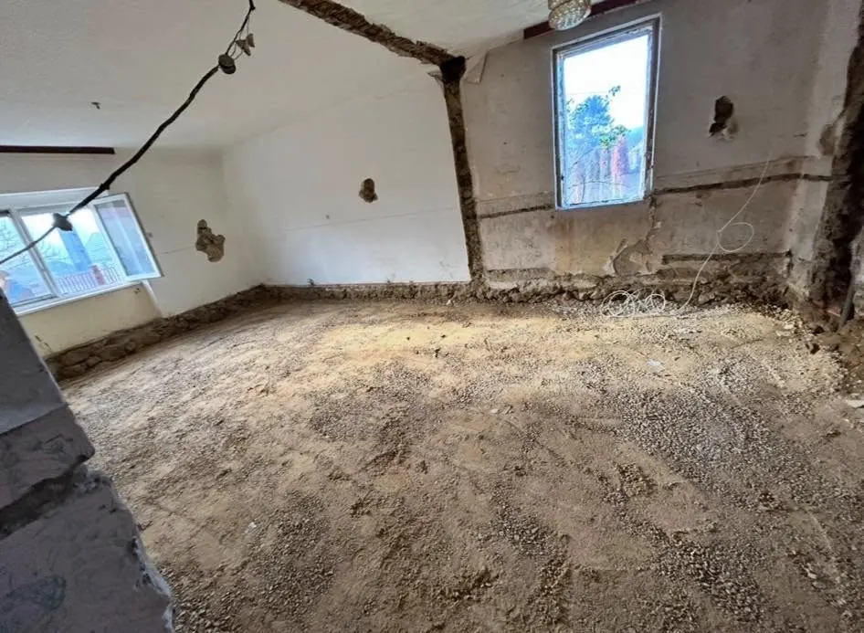 A kép egy Kőszegen található generálkivitelezési projekt helyszínét mutatja be, ahol a falak részben kibővítve, a padló pedig még befejezésre vár. Az üres teret por és homok borítja, míg az ablakok mögött természetes fény világítja meg a teret. A Farkas-Konstrukt Kft. szakértelme nyomán a helyszín átalakulásra készül.