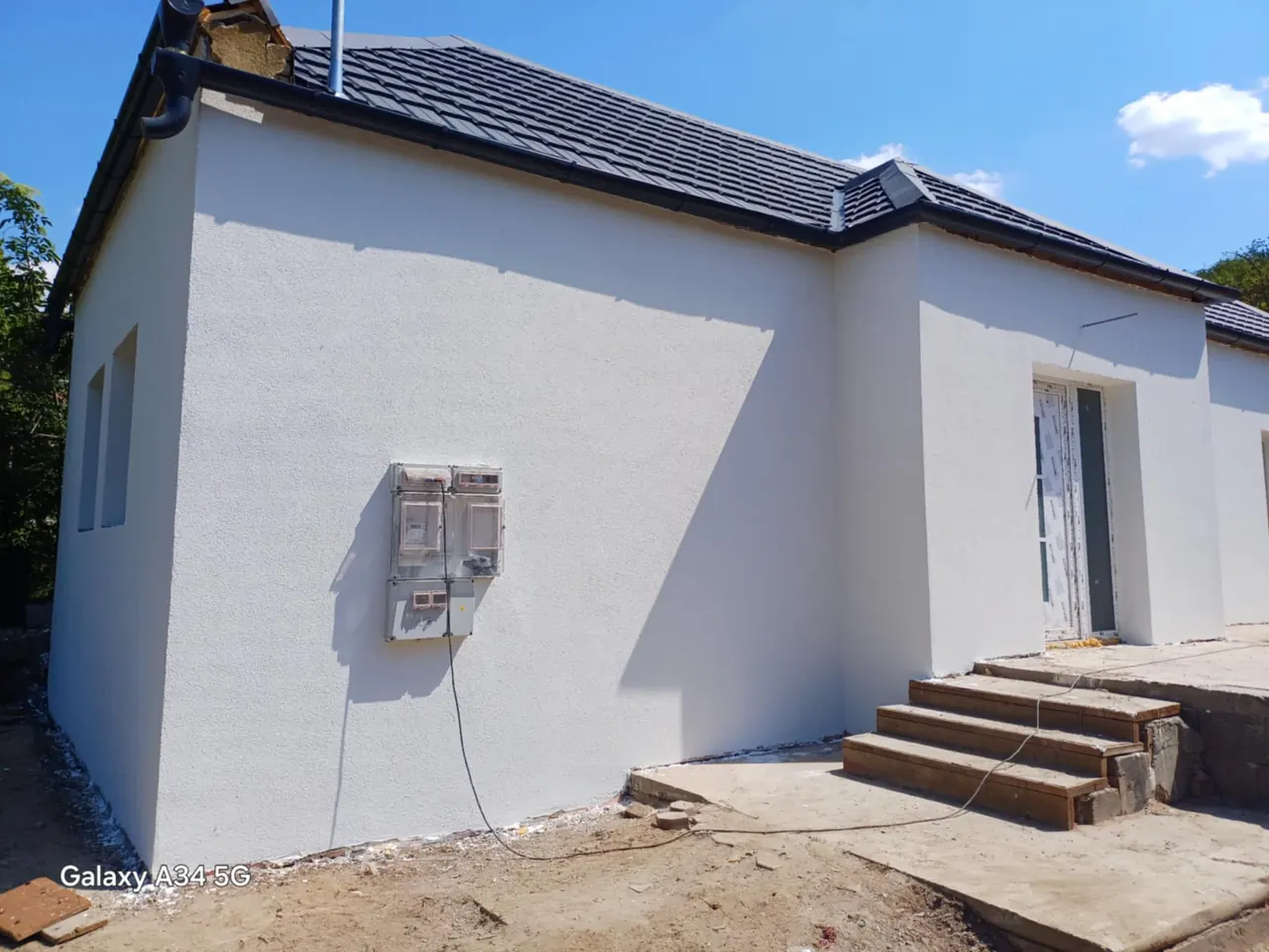 A képen egy frissen épített ház látható, amelynek sima, fehér falai és modern tetőszerkezete van. A lépcső vezet a bejárati ajtóhoz, amely még nincs befejezve. Az épület mellett elektromos mérőórák találhatók. A munka Győr városában zajlik, ahol a Farkas-Konstrukt Kft. generálkivitelezési szolgáltatásait nyújtja.