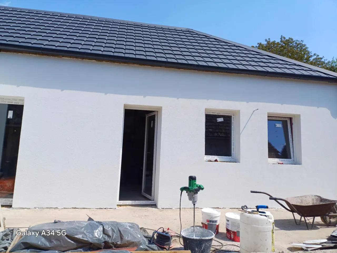 A képen egy frissen épült ház látható Csornán, ahol a Farkas-Konstrukt Kft. generálkivitelezési munkálatai zajlanak. Az épület sima, fehér falakkal és fekete tetőcseréppel rendelkezik. Az építkezés területén különböző szerszámok és anyagok, például festékek és betonkeverő láthatók, amelyek a kivitelezéshez szükségesek. A háttérben kék égbolt figyelhető meg.