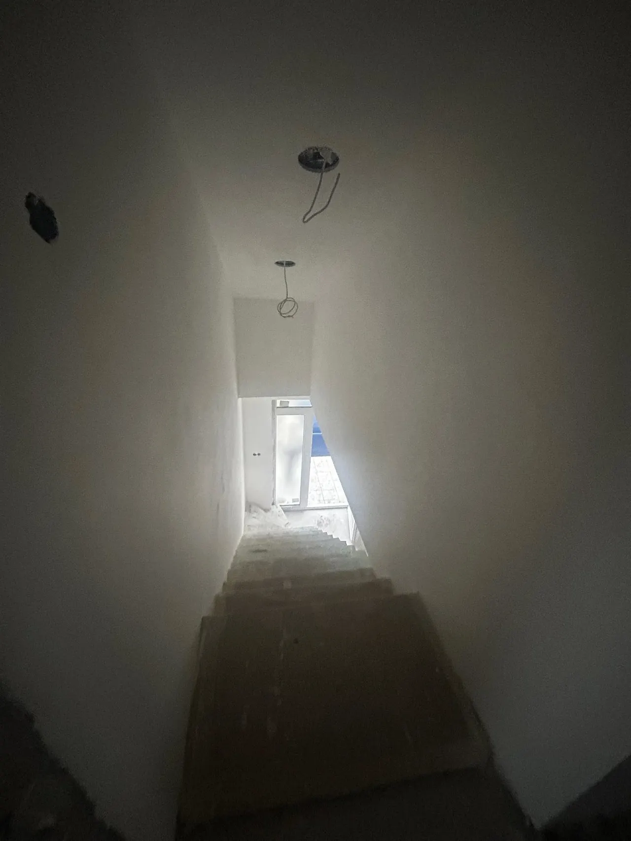 A képen egy építkezés alatt álló lépcsőház látható, amely Győr városában található. A falak fehérre meszelve, a lépcsők burkolás előtt állnak. A háttérben egy ajtó nyílik, amelyen át természetes fény áramlik be, hangsúlyozva a Generálkivitelezés során végzett munkát. A helyiség minimalista stílusú, a munkálatok még folyamatban vannak.