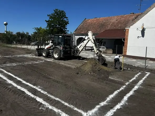 Győr városában a Farkas-Konstrukt Kft. generálkivitelezési szolgáltatásai keretében földmunka folyik. A képen egy munkagép látható, amint éppen földet ás és formál a területen, amelyet fehér vonalakkal határoltak. A háttérben egy épület és zöld fák találhatók, így jól érzékelhető a helyszín környezete.