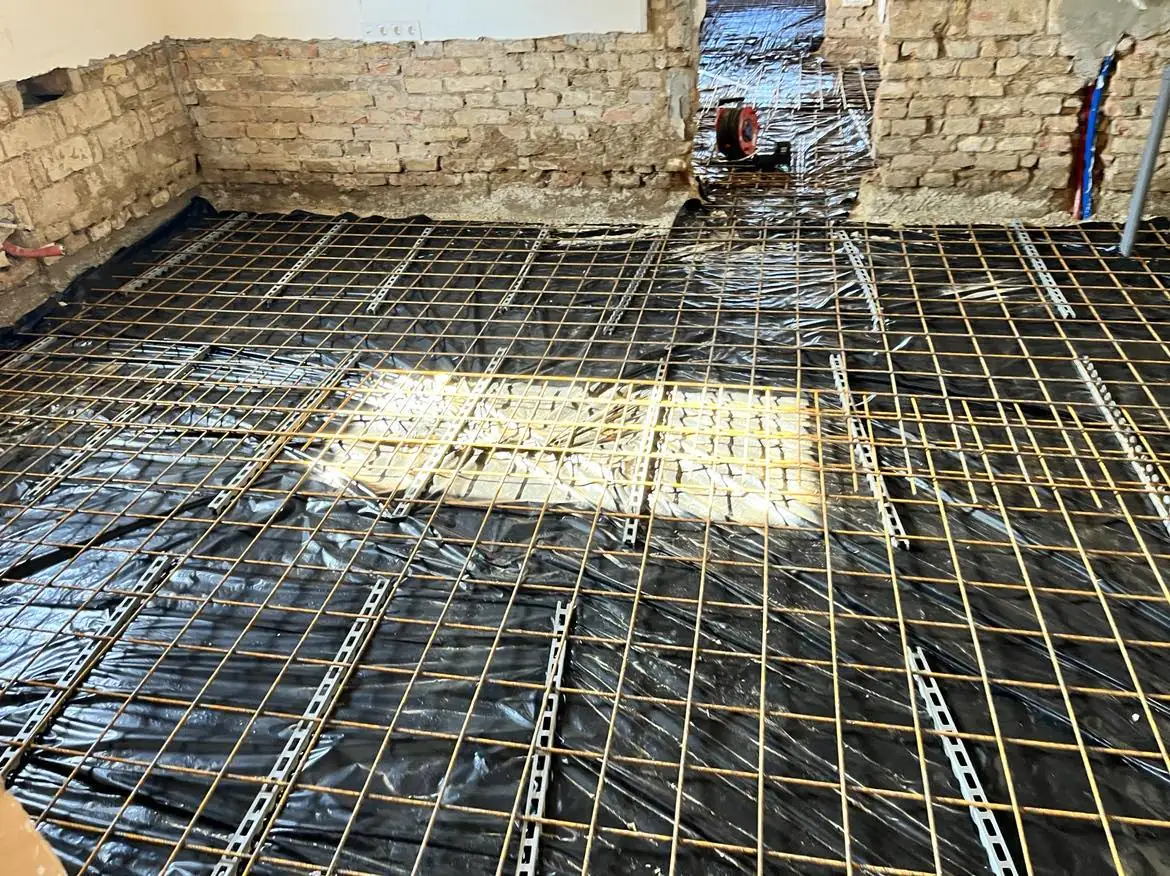 A képen egy építkezési terület látható, ahol a padló alatti alapozás folyamatban van. A falak téglából készültek, és a padlón fém rácsozat található, amely a Generálkivitelezés munkálatait tükrözi. A háttérben egy nyitott ajtó látható, ami egy másik helyiségbe vezet, míg a Győr városában lévő projekt előrehaladását jelzi a szakszerű kivitelezés.