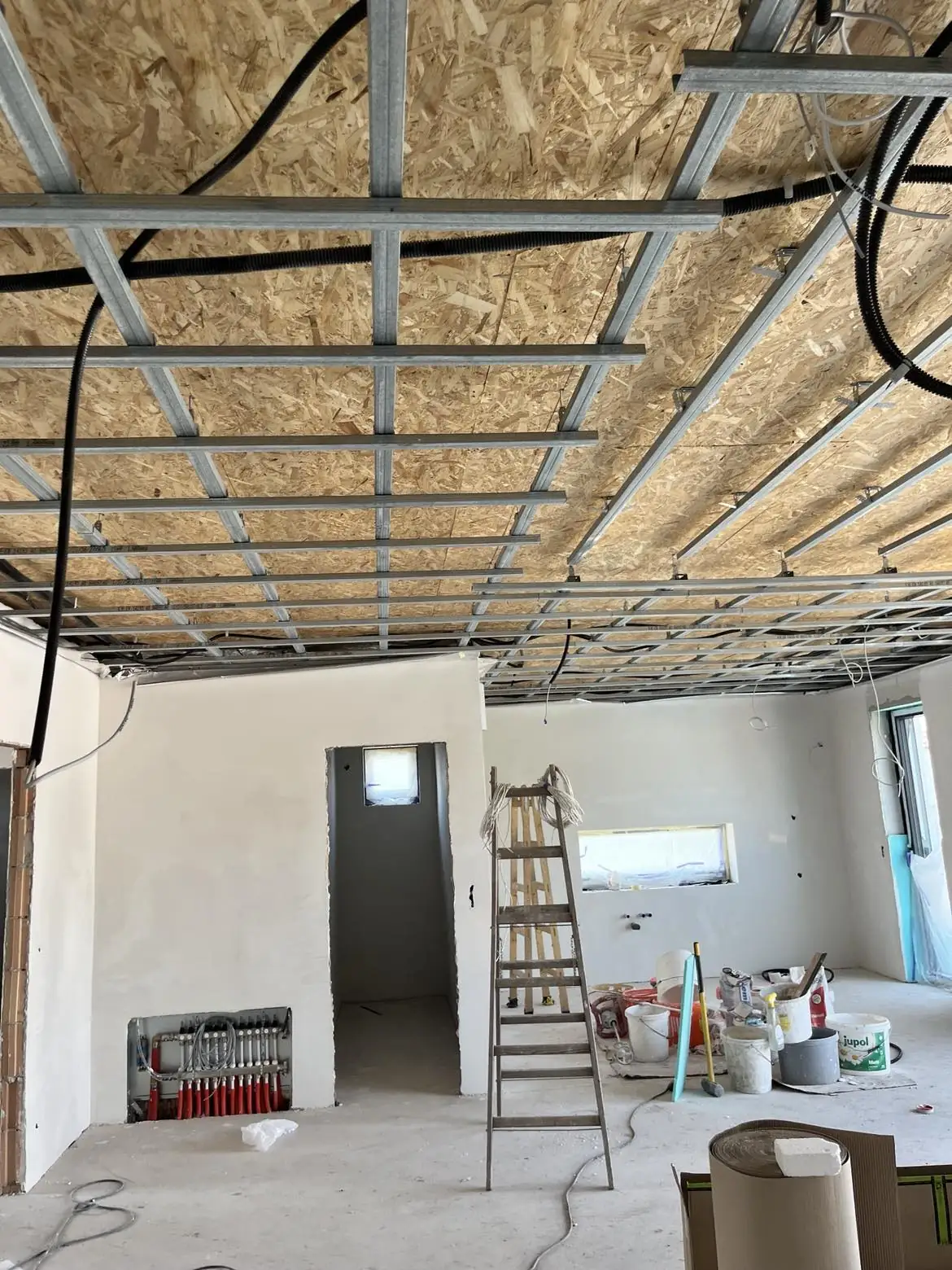 A képen egy építkezés látható, ahol a plafon szerkezete fémprofilokból készült, és az épület belső terében dolgoznak. A falak fehérre vannak meszelve, és a padlón különböző építőanyagok, valamint egy létra található. A háttérben látszik a bejárati ajtó és a fűtéscsövek. Ez a helyszín Sárváron található, ahol a Farkas-Konstrukt Kft. Generálkivitelezés szolgáltatását végzi.