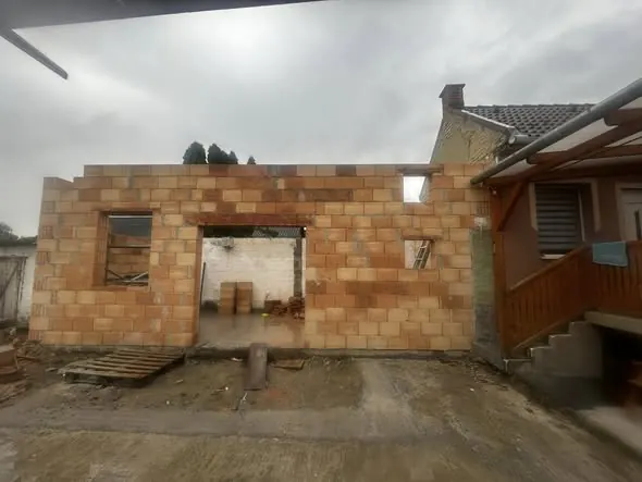 A képen egy építkezés látható Győr városában, ahol a Farkas-Konstrukt Kft. generálkivitelezési munkálatai zajlanak. Az épület falai már burkolva vannak, és a tető zöld színű cseréppel van fedve. A háttérben egy fehér furgon és egy mobil vécé látható, míg az udvaron építőanyagok és munkaeszközök találhatók. Az idő borongós, ami a munkálatok előrehaladását jelzi.