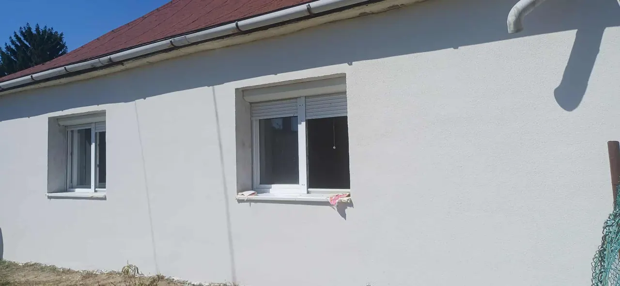 A képen egy frissen felújított házfal látható, amely Győr városában található. A fal sima, fehérre festett, két ablak látható rajta, mindkettő zárva van. A tető piros cserépborítással készült. A Farkas-Konstrukt Kft. által végzett generálkivitelezés munkálatai jól tükröződnek a ház állapotában, amely a modern építkezés színvonalát képviseli.