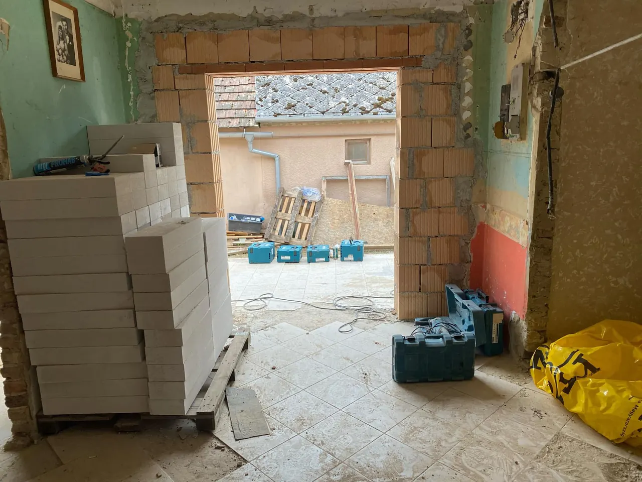 A képen egy építkezés látható, ahol a Farkas-Konstrukt Kft. Generálkivitelezés szolgáltatása keretében családi ház tervezése zajlik Győr városában. Az előtérben fehér téglák halmozva, míg a háttérben egy nyitott ajtó vezet a szabadba, ahol építőanyagok és szerszámok találhatók. A falak még befejezésre várnak, a padló burkolata pedig félig kész.