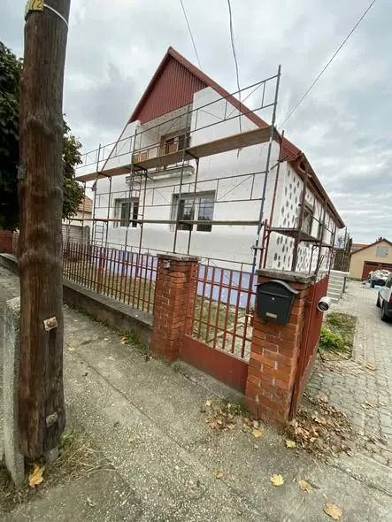 A képen egy építés alatt álló ház látható Győrben, ahol a Farkas-Konstrukt Kft. generálkivitelezési szolgáltatását végzik. Az épületet állványzat veszi körül, és a falak fehér színűre vannak festve. A kerítés mögött látható a ház, amely kulcsrakész kivitelezés részeként készül. Az utcában parkoló autók és zöldellő fák találhatóak, amely a városban jellemző környezetet tükrözi.