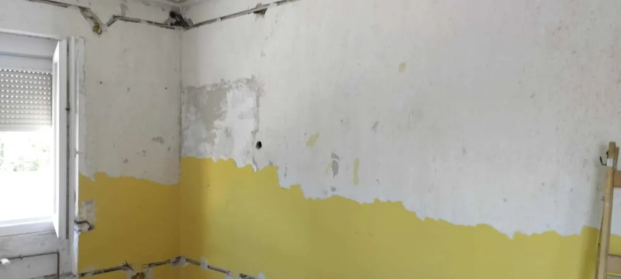 A képen egy felújítás alatt álló szoba látható, ahol a falak részben sárga színűre vannak festve, míg más részek még festésre várnak. A szoba ablakánál látható egy nyitott redőny, amelyen át beáramlik a természetes fény. A kép Győr városában készült, a Farkas-Konstrukt Kft. Generálkivitelezés projektje keretében.
