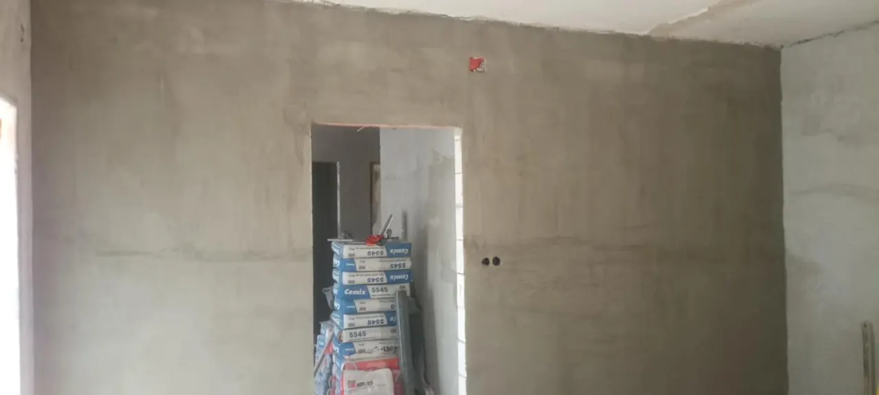 A képen egy frissen vakolt fal látható, amely a Sopronban található Farkas-Konstrukt Kft. generálkivitelezési projektjéhez tartozik. A fal mellett egy ajtó nyílása is látható, amelyen keresztül a következő helyiségbe lehet belépni. A földön építőanyagok találhatók, amelyek a kivitelezési munkálatokhoz szükségesek.