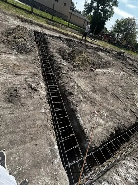 A képen a Farkas-Konstrukt Kft. generálkivitelezési munkálatai láthatók Győrben. A földmunkák során kialakított alapozás látható, amelyben acélszerkezet helyezkedik el. A háttérben munkások dolgoznak a területen, amely még készülőben van. A terület körül fák és épületek találhatók, ami a helyszín természetes környezetét tükrözi.