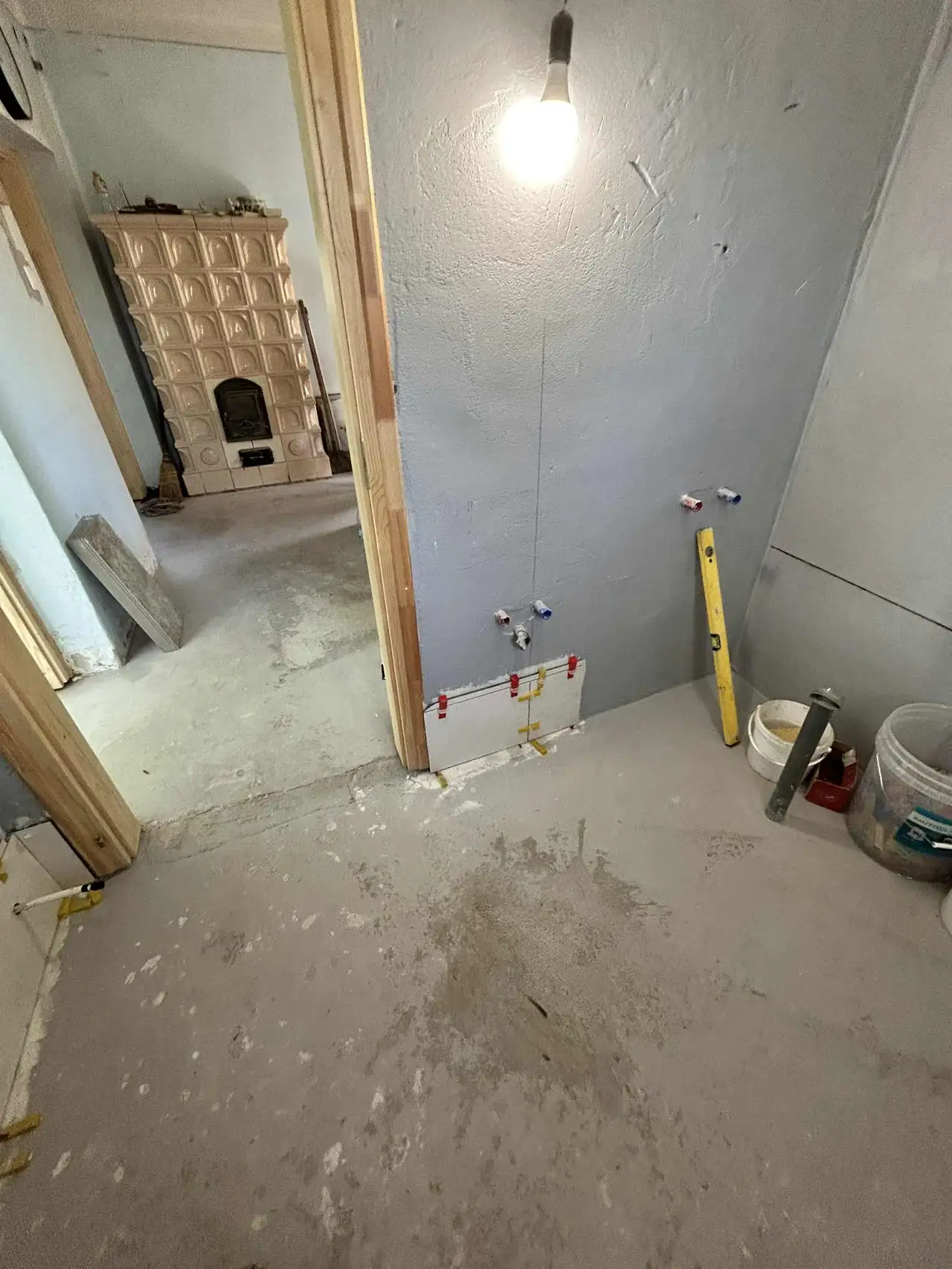 A képen egy fürdőszoba felújításának előkészítése látható, ahol a falon vízcsövek kiállásai és a szükséges szerelvények láthatók. A helyiségben világító lámpa van, amely megvilágítja a munkaterületet. A padló betonozott, és a falak szürke színűek. Az ilyen generálkivitelezési munkák jellemzőek a Farkas-Konstrukt Kft.-nél Győr városában.