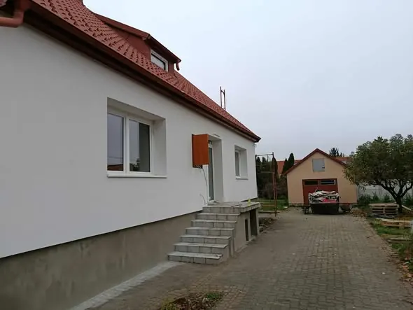 Győr városában található Farkas-Konstrukt Kft. generálkivitelezési projektje látható, amely egy modern, világos színű ház oldalát mutatja be. Az épülethez lépcső vezet, és a területen egy másik épület is látható a háttérben. A környezet rendezett, a kertben fák és egy rakodóhely található, ami hangsúlyozza a cég szakmai hozzáértését.