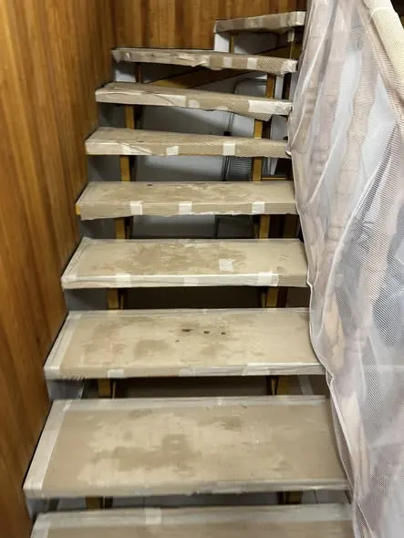 Az Ajkán található Farkas-Konstrukt Kft. generálkivitelezés projektjének lépcsője látható az imágon, amelyen a lépcsőfokok védőfóliával borítva állnak. A lépcsőfokok fából és betonból készültek, a háttérben pedig fa burkolat látható, ami meleg hangulatot kölcsönöz a térnek. A lépcsőház biztonságos használatát szolgáló tűződrótok is megfigyelhetők.