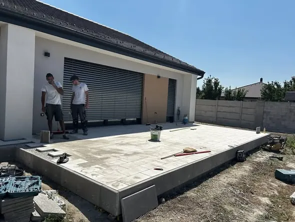 Két munkás dolgozik egy ház teraszának burkolásán, Győr városában. Az új burkolat szürke járólapokból készült, és a terasz előtt helyezkedik el. A háttérben látható a ház falazata és a redőny, míg az előtérben szerszámok és anyagok sorakoznak. A Farkas-Konstrukt Kft. generálkivitelezésének részeként a munkálatok a terasz kialakítását célozzák.