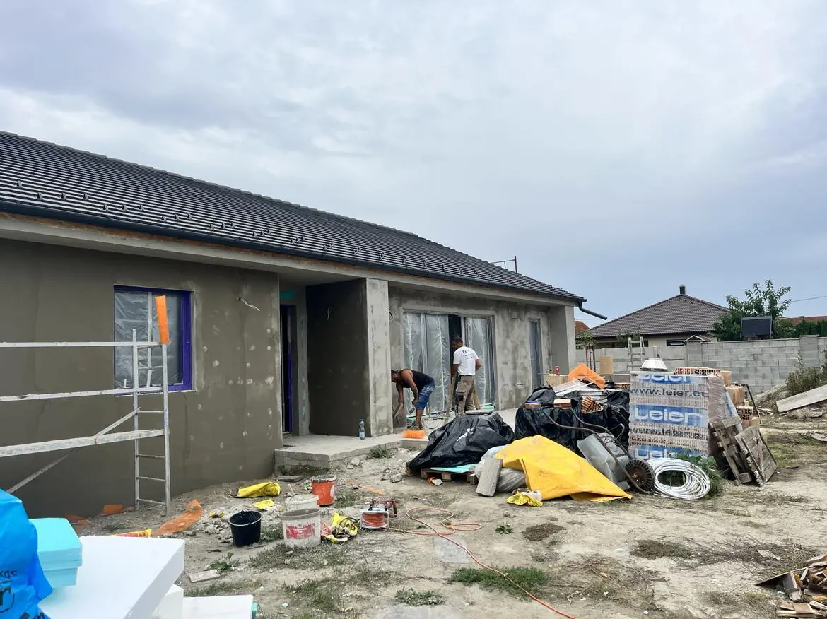 Győr városában, a Farkas-Konstrukt Kft. generálkivitelezési projektjén egy családi ház építkezése látható. A képen a munkások a ház külső falán dolgoznak, miközben a területen építőanyagok, mint például cement, zsákok és különböző szerszámok találhatóak. A háttérben a ház már részben kész, a tető és az ablakok kialakítása jól látható.