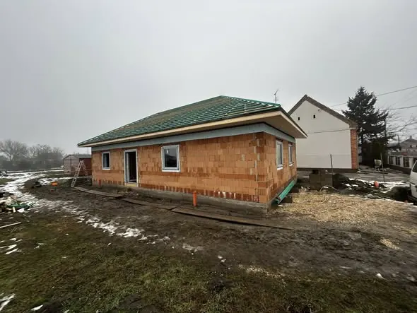 Farkas-Konstrukt Kft. generálkivitelezési projektje látható Győrben, ahol egy új ház építkezése zajlik. A képen a frissen falazott ház látható, zöld tetővel és nagy ablakokkal, körülötte építkezési terület és fűtlen zöld terület. Az idő borongós, de a projekt előrehaladása jól látható.