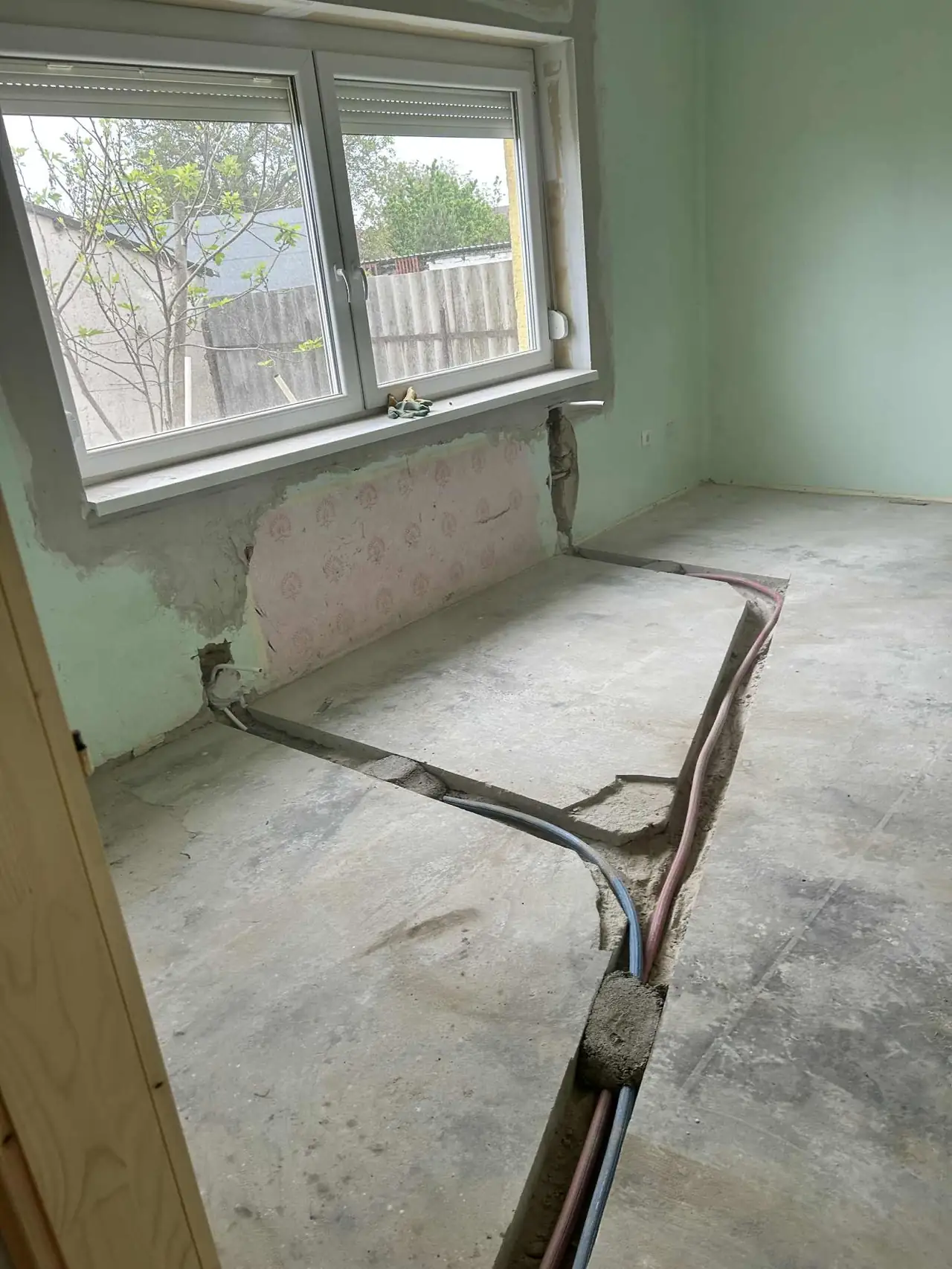 A képen egy még be nem fejezett belső tér látható, amely Győr városában található. A falak zöldre festettek, és a padló burkolata le van bontva, ami a generálkivitelezés folyamatát jelzi. A falból különböző vezetékek és csövek látszanak ki, amelyek a jövőbeli elektromos és vízvezeték-rendszer részei lesznek. A Farkas-Konstrukt Kft. szakértelme nyomán a munka előrehalad.