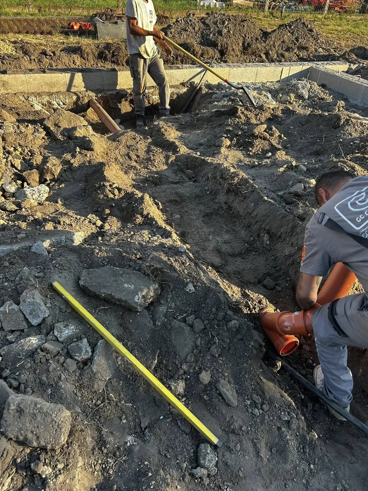 Két munkás a Farkas-Konstrukt Kft. földmunkáit végzi Győrben, ahol a Generálkivitelezés keretében alapozási feladatokat hajtanak végre. Az egyik munkás ásóval dolgozik, míg a másik egy csőcsatlakozást helyez el a frissen ásott gödörben. A háttérben építőipari gépek és berendezések láthatók, amelyek a kivitelezési munkálatokat segítik.