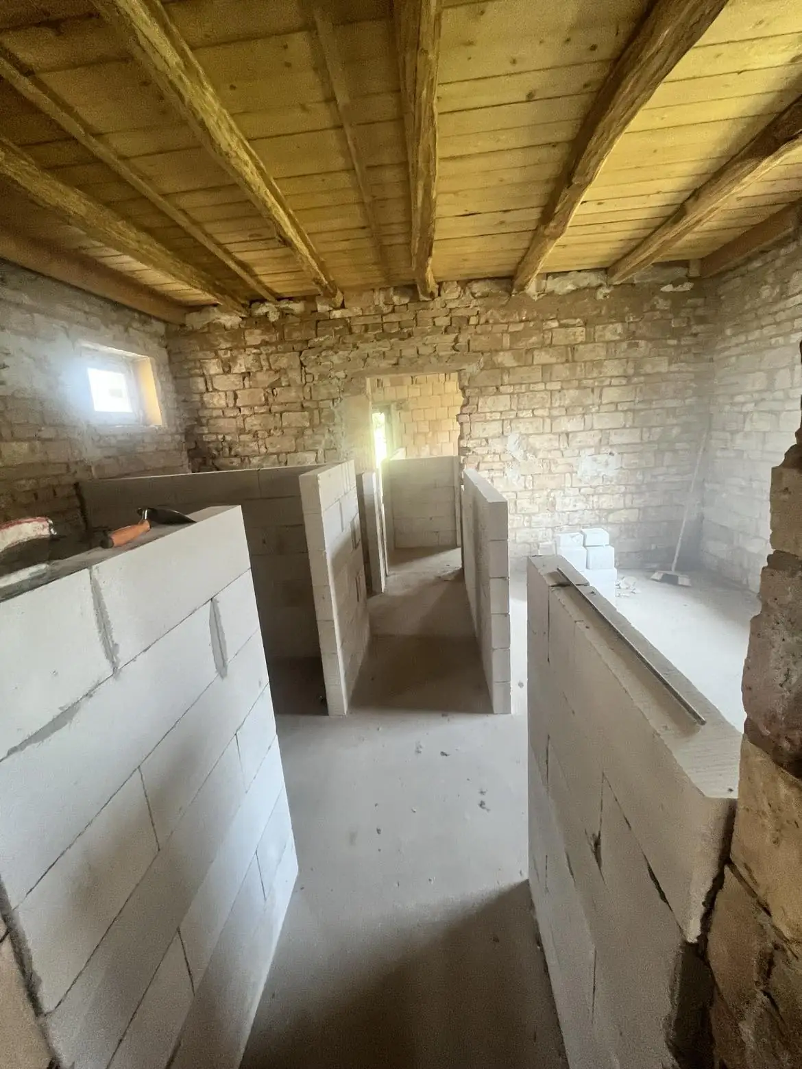A képen egy építkezés látható, ahol a falak már részben ki vannak alakítva. A helyiség falai szürke téglákból készültek, a mennyezet fából van. A világítás gyenge, de a napfény szűrődik be az ablakokon. Csornában, a Farkas-Konstrukt Kft. generálkivitelezési projektjén készült felvétel, amely a belső terek kialakítását mutatja.