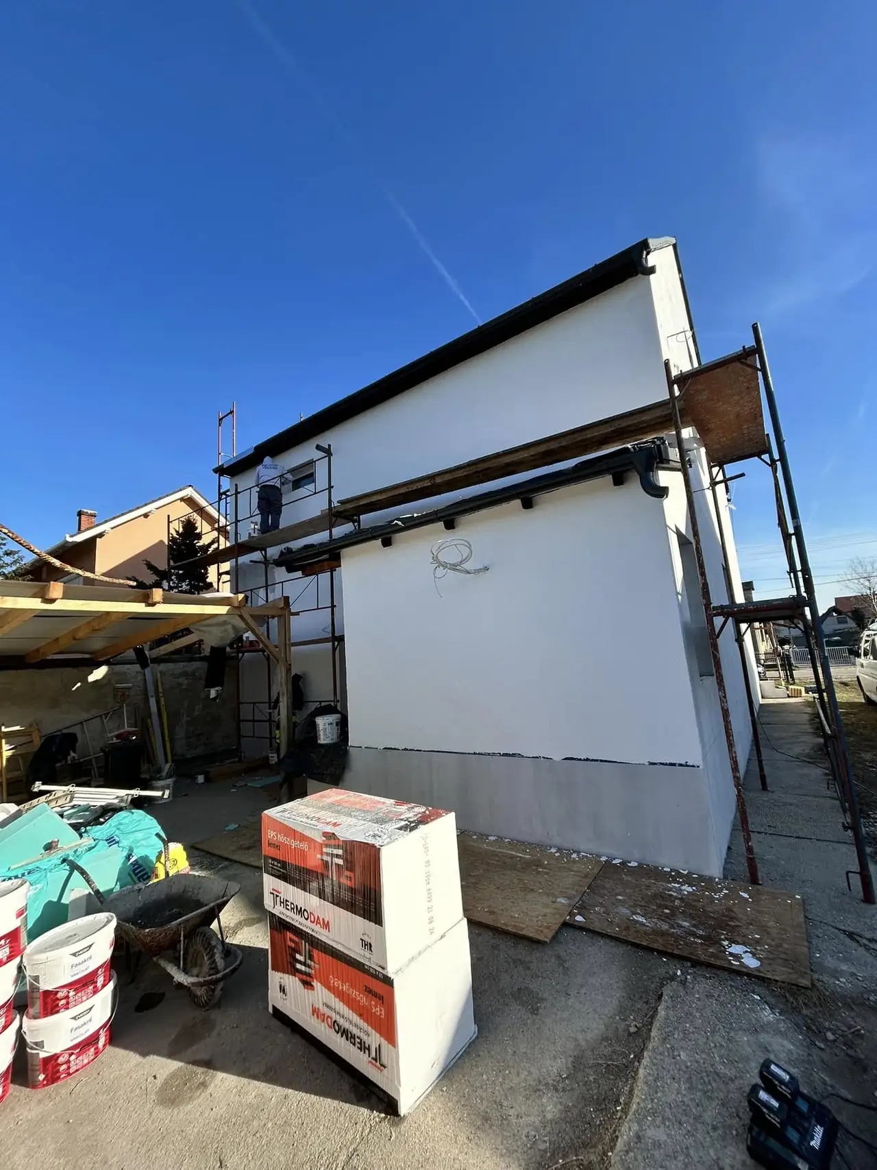 Az építkezés folyamatát bemutató képen egy frissen vakolt fal látható, amely mellett állványzat és építőanyagok találhatók. A háttérben más épületek is megfigyelhetők, míg a területen különböző szerszámok és anyagok helyezkednek el. Ez a kép a Farkas-Konstrukt Kft. generálkivitelezési munkálatait illusztrálja Győr városában.